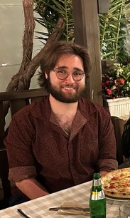 George, a white male with medium-length brown hair and a beard, smiles at the camera. He is sitting at a table with a pizza and green bottle in front. He is wearing a brown button-up shirt with the sleeves rolled up to his elbows and brown round glasses.