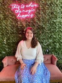 Alexis, a white woman with long brown hair, smiles at the camera as she sits on a pink couch in front of a leaf-covered wall. 