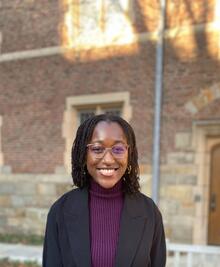 Chisom, a Black woman with shoulder-length black mini twists, smiles at the camera. She is wearing gold-rimmed glasses and gold hoops. She has on a dark purple turtleneck and a black blazer. There is a brick building in the background.&nbsp;